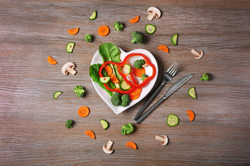 Useful cut vegetables on a plate in the form of heart on wooden table top view
