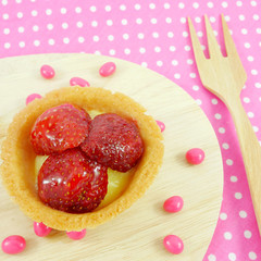 The delicious home baked strawberry tart and small pink chocolate candies on the wooden board with pink cotton fabric.