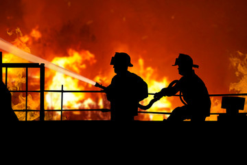 Пожарные тушат огонь на крыше дома. Силуэты пожарных на фоне пожара, огня. Двое мужчин в форме пожарных тушат огонь на крыше дома из брансбойта
