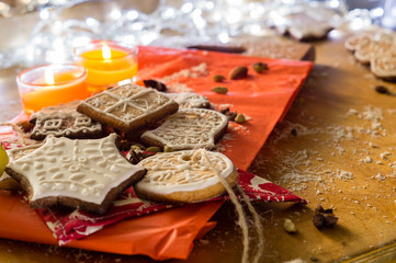 Christmas ginger cookies covered with white icing, almonds and spices on a light brown wooden background and red paper stand. They are illuminated by white lights and candles.