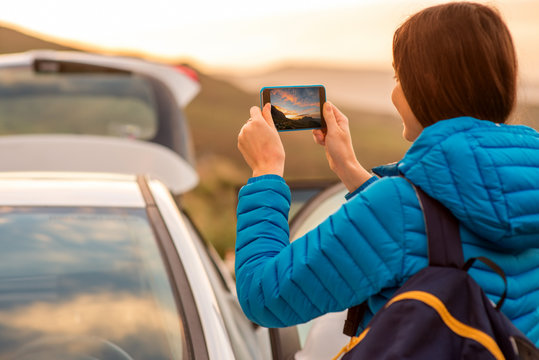 Female traveler photographing sunrise with smartphon near the car