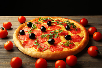 Tasty pizza with salami and olives decorated with tomatoes on wooden table against black background
