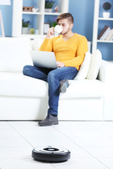 Cleaning concept - automatic robotic hoover clean the room while man relaxing with laptop, close up
