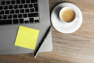 Empty yellow adhesive paper on laptop keyboard, pen and coffee cup on desk background