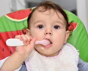 Smeared hungry baby girl eating baby food with plastic spoon