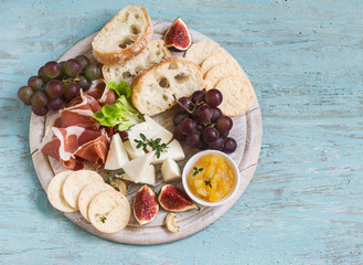 delicious appetizer to wine - ham, cheese, grapes, crackers, figs, nuts, jam, served on a light wooden board, and two glasses with white wine on bright wooden surface