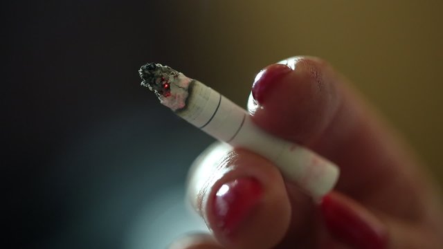 A senior woman hand with cigarette, female smoker, close up shot. Smoldering cigarette in  woman hand, lady with red nails, cigarette smoke flows up and down, beautiful manicure, bright nail polish