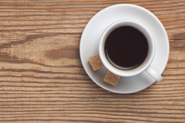 White saucer and cup of coffee with two cane sugar pieces on rustic wooden table background top view have place for text