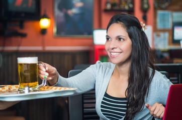 Brunette model sitting by restaurant table holding glass of beer and posing with positive attitude smiling