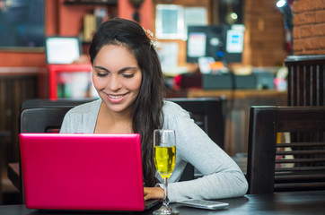 Attractive brunette wearing grey sweater sitting at restaurant table working with pink laptop and glass of champagne
