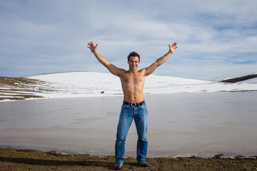 Uomo a torso nudo e braccia alzate in riva a un lago ghiacciato. Sfondo con montagne innevate