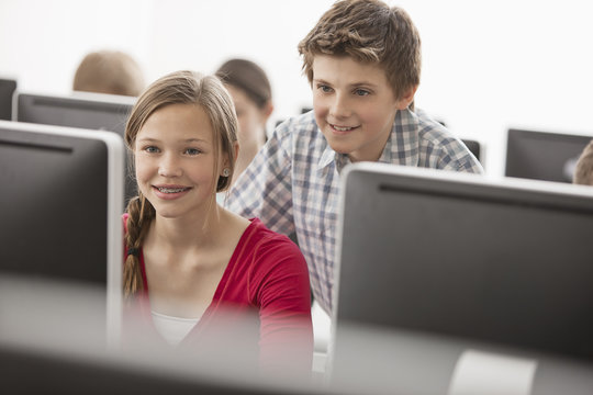 A school computer lab room, with rows of screens. Two young people looking intently at the screen.