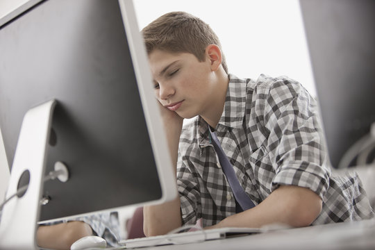A school room, computer laboratory or lab with rows of computer monitors and seating. A boy sitting with his hand on his chin looking fed up. 