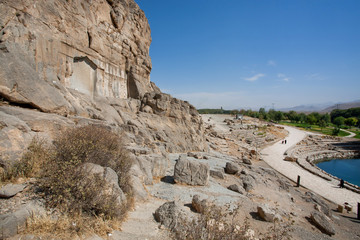 Rocks over the lake and ancient stone reliefs in the persian valley with rural road, Iran