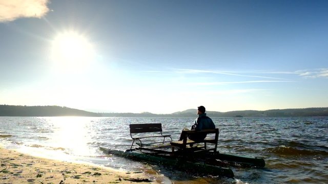 Tourist in blue jacket sit on abandoned old rusty paddle boat stuck on sand of beach. Wavy water level, island on horizon. Autumn weather at coastline 