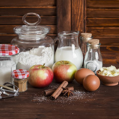 Ingredients and tools for baking - flour, eggs, butter, apples, cinnamon on a brown rustic wooden surface.  Ingredients for baking apple pie