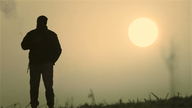 The man smokes a cigarette at dawn. A young man in a field at dawn, with a cigarette in his hand. Bearded man at dawn.