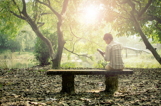 Young man relaxing & reading books alone in a peaceful garden enjoying his leisure in early winter afternoon