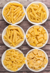 Various dried pasta variety and shapes in white bowl over wooden background