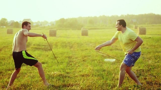 Young friends play the fool in the field during sunset with wooden rapier in slowmotion