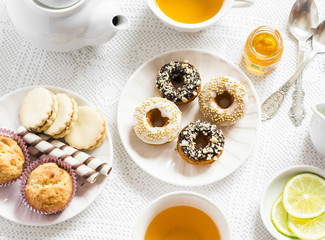Lemon green tea and sweets - banana muffins, cookies with caramel and nuts, donuts with chocolate and lemon glaze, tea set on white table cloths on a light surface. Tea time. Vintage and rustic style
