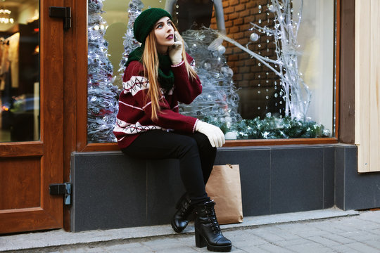 Street portrait  of fashionable beautiful young woman sitting near the shop-window and speaking on mobile phone. Lady wearing stylish winter clothes. Female fashion concept. 