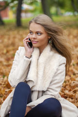 Young happy blonde woman calling by phone in autumn park