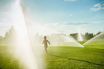 A little boy running around the green fields under the spray of fountains.