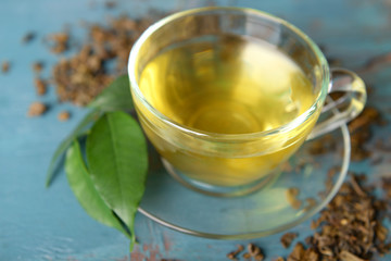 Glass cup of tea with green leaves and scattered tea around on blue wooden background