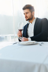 Drink. Closeup Of Elegant Handsome Young Man Drinking Red Wine Sitting In Luxury Gourmet Restaurant. Businessman Relaxing And Looking Through The Window. Success Celebration Dinner Concept