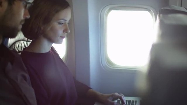 Young woman is working on a laptop inside an airplane next to a window while a man talks to her. Shot on RED Cinema Camera.