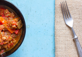 Vegetarian dish of stewed vegetables in a black ceramic bowl and fork in stainless steel on canvas on old blue table. The view from the top. Rustic style. Free space for text. Copy space