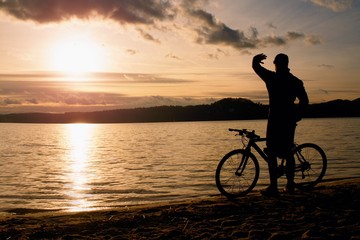 Obraz premium Young man cyclist silhouette on blue sky and sunset above the beach. Biker atthe end of season at lake.