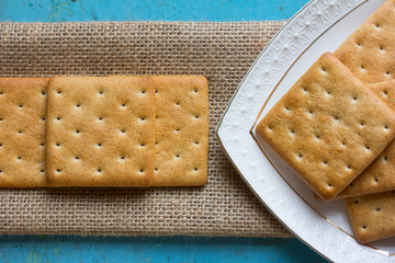 Square cracker biscuits on a linen napkin and a triangular portion of white ceramic plates with cookies on old blue table. Close up, top view