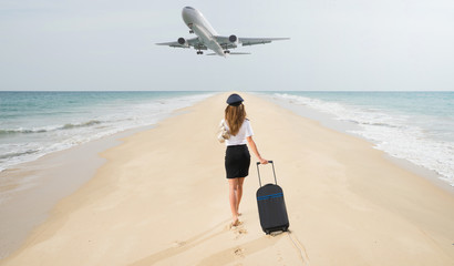 Travel concept. Young woman in flight attendant clothes walking on the beach with suitcase and hat . Overhead fly plane. 