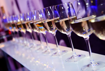 Nightclub wine glasses with white wine lit by party festive lights on dark-purple background,...
