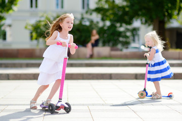 Two adorable little sisters riding their scooters in a summer park