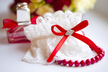 Подвязка невесты из белого кружева с красной лентой и камнями на фоне красных бус и духов