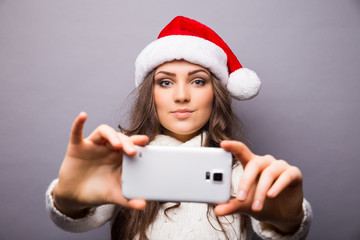 Christmas portrait of beautiful girl. Girl wearing santa claus hat. Girl smiling and making selfie on mobile phone