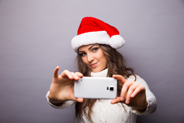 Christmas portrait of beautiful girl. Girl wearing santa claus hat. Girl smiling and making selfie on mobile phone