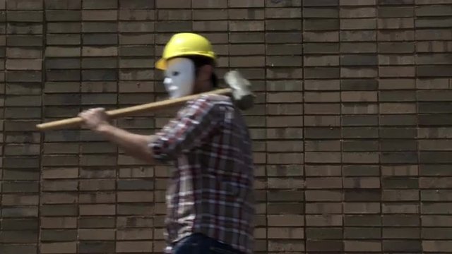 Two masked contractors wearing hard hats, while carrying a sledgehammer and an adjustable wrench, walk endlessly past the same brick wall.  Locked off wide shot.  Looping clip.