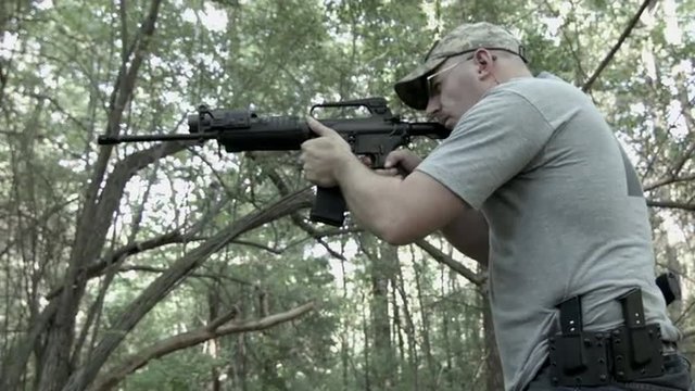 Profile mid shot of man firing an AR-15 assault rifle in woodland setting.  Originally recorded in 4K, Ultra High Definition.