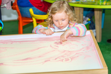 Fototapeta na wymiar Cute child girl drawing draws developing sand in preschool at table in kindergarten by method of Montessori
