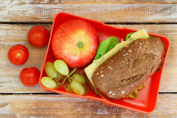 Healthy school lunch box containing whole grain roll with cheese and lettuce, cherry tomatoes, red apple and grapes 

