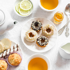 Lemon green tea and sweets - banana muffins, cookies with caramel and nuts, donuts with chocolate and lemon glaze, tea set on white table cloths on a light surface. Tea time. Vintage and rustic style