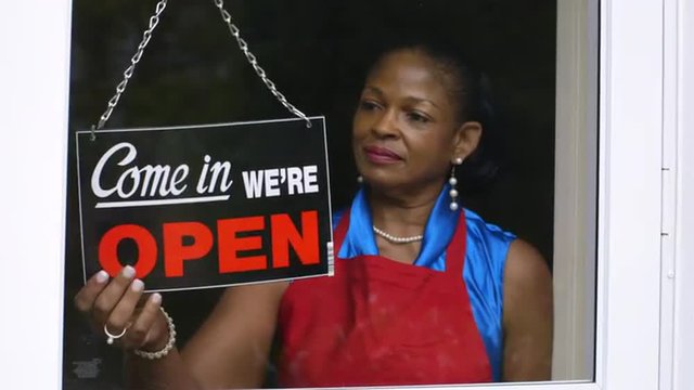 A lovely African American woman changes the window sign on her small business from closed to open then smiles a great smile.