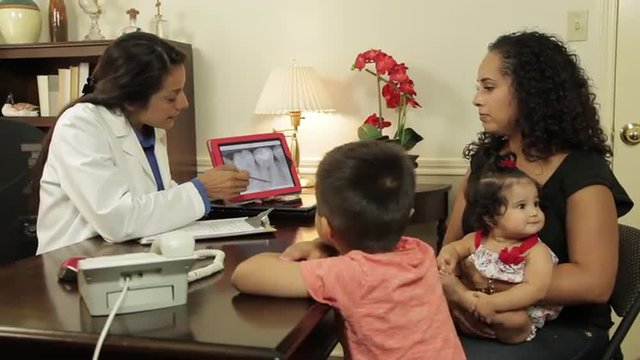 A Hispanic dentist in her office with a mom and her two children going over dental x-ray image displayed on a tablet pc.