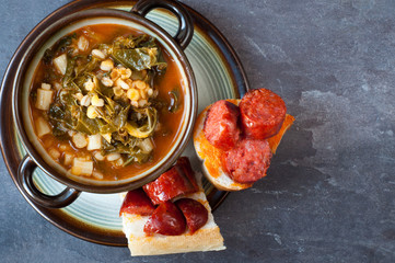 Kale, pearl barley and tomato soup served with crusty bread topped with chunks of chorizo sausage. Presented in a rustic bowl on a slate stone table.