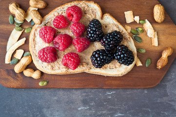 Fototapeta premium Healthy snack of sliced sourdough bread topped with peanut butter and fresh blackberries and raspberries. Served on a wooden tray on a slate stone table top.