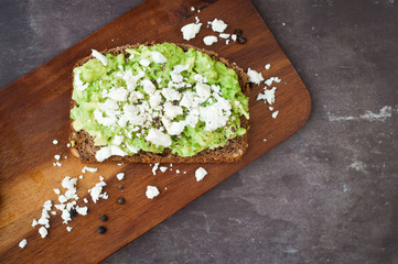 Healthy snack of dark rye bread topped with avocado and crumbled feta cheese served on a stone slate table top.